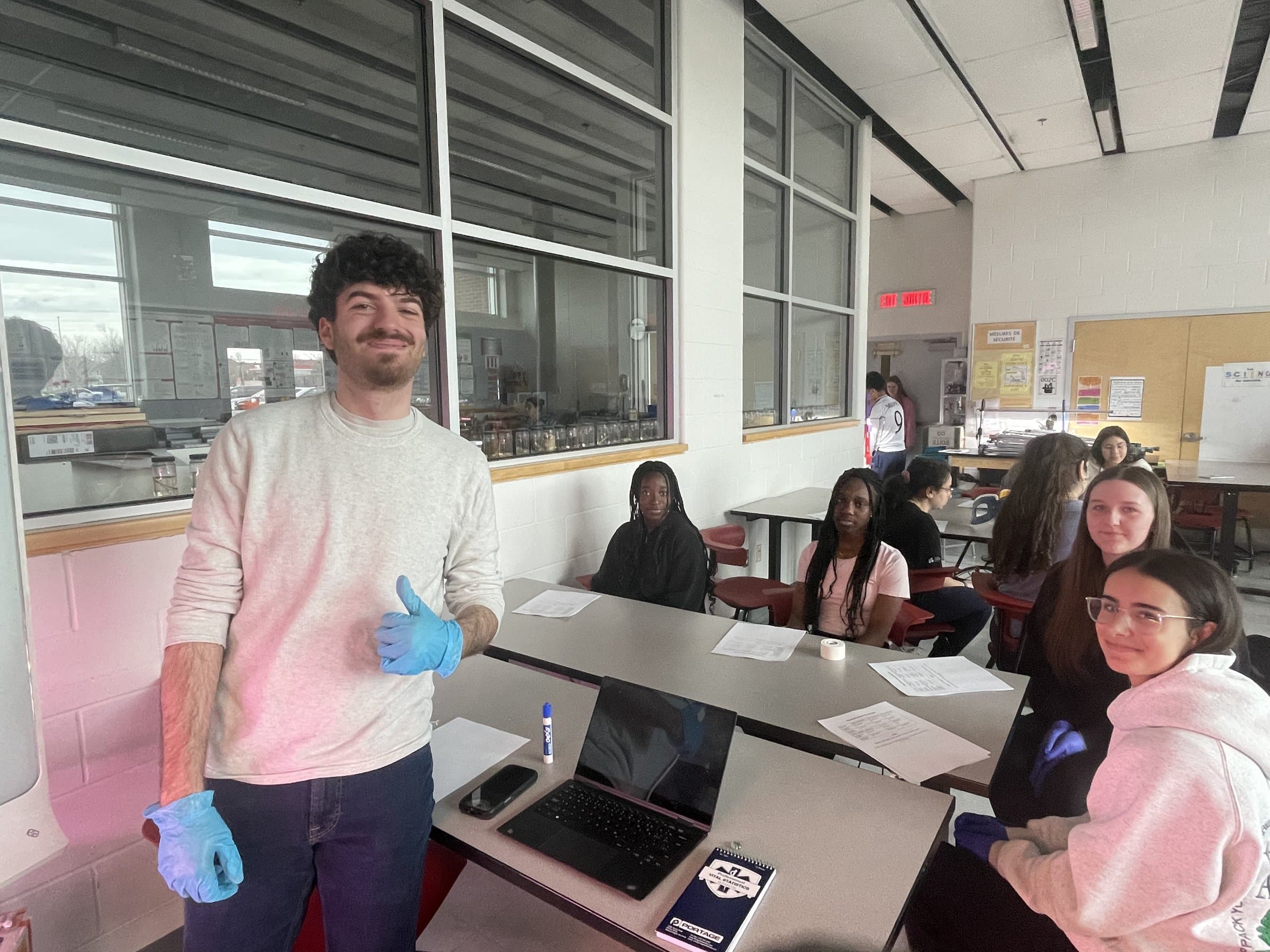 A mentor in blue medical gloves gives a thumbs up while students look on from their tables