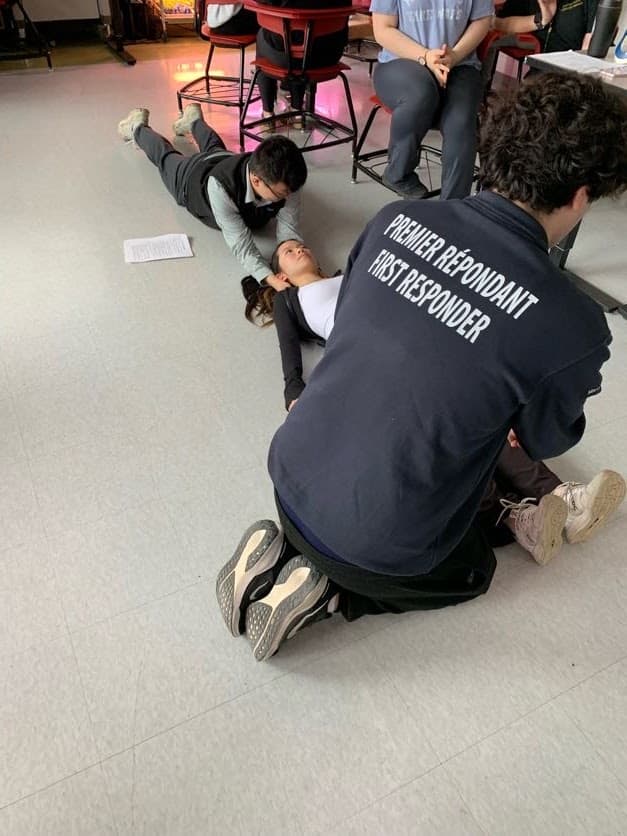 Two students kneel beside classmates acting as patients during a first aid simulation